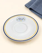 Detalhe do pires branco Family Cook com aros azul e amarelo e frase sabor em família